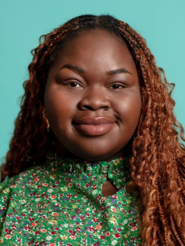 Portrait of a person with long, curly hair, wearing a green floral top, smiling slightly against a turquoise background.